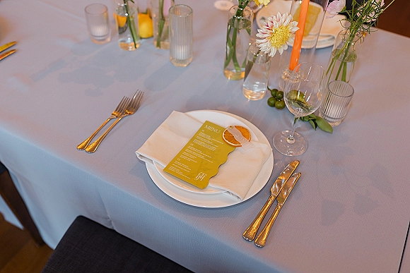 Reception tablescape with wedding place setting, gold flatware, menu card on white napkin, dried orange slice and taper candle on white tablecloth
