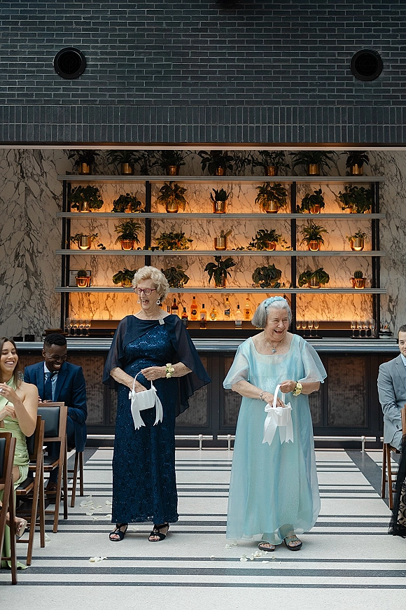 Wedding ceremony moment with flower girls tossing petals, elderly women in formal gowns carrying ribbon baskets down a striped indoor aisle