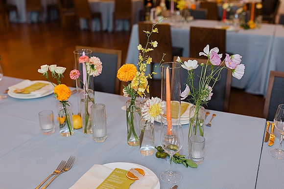 Reception tablescape with bud vase centerpiece, pastel taper candles and tall glass cylinders on blue linen, gold flatware and citrus place cards on set banquet tables
