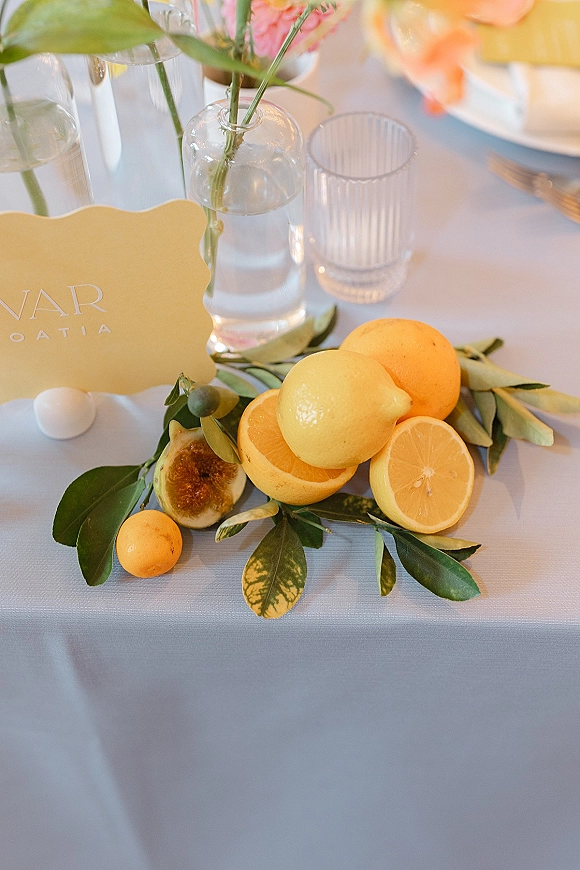 Reception tablescape with a citrus wedding centerpiece of lemon slices, oranges, figs and greenery, plus pink bud vases and ribbed votives on white linen