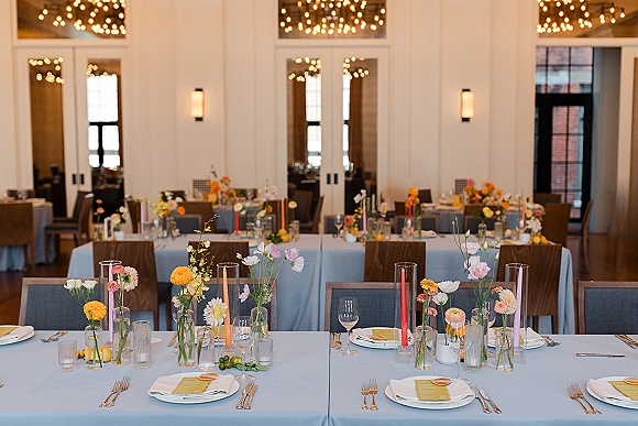 Reception tablescape with wedding bud vase centerpieces, pastel taper candles, and gold flatware on light blue linens in a modern banquet hall with chandeliers