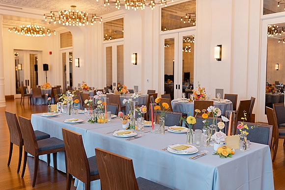 Reception tablescape with light blue tablecloth, bud vase florals, orange taper candles, citrus accents on a long banquet table in a modern ballroom