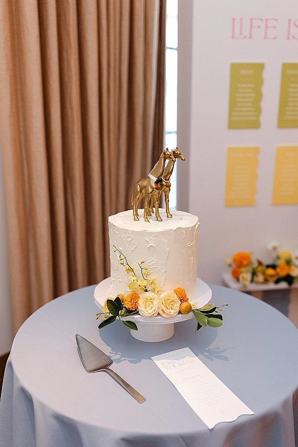 Wedding cake with textured buttercream, gold giraffe topper, and rose and orange floral cluster on a blue tablecloth by beige curtains