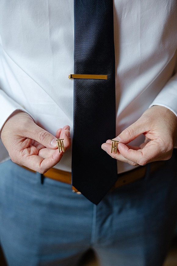 Groom accessories with wedding cufflinks in gold, paired with a gold tie clip on a black necktie over a white shirt and blue trousers
