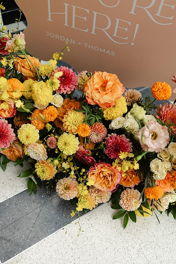 Wedding welcome sign with flowers, arched printed board on a stone floor, grounded with dahlias, roses, marigolds and greenery