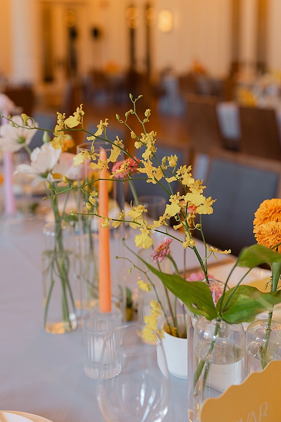 Reception tablescape with wedding bud vase centerpiece of yellow, orange, and pink blooms alongside taper candles and glassware in warm lighting