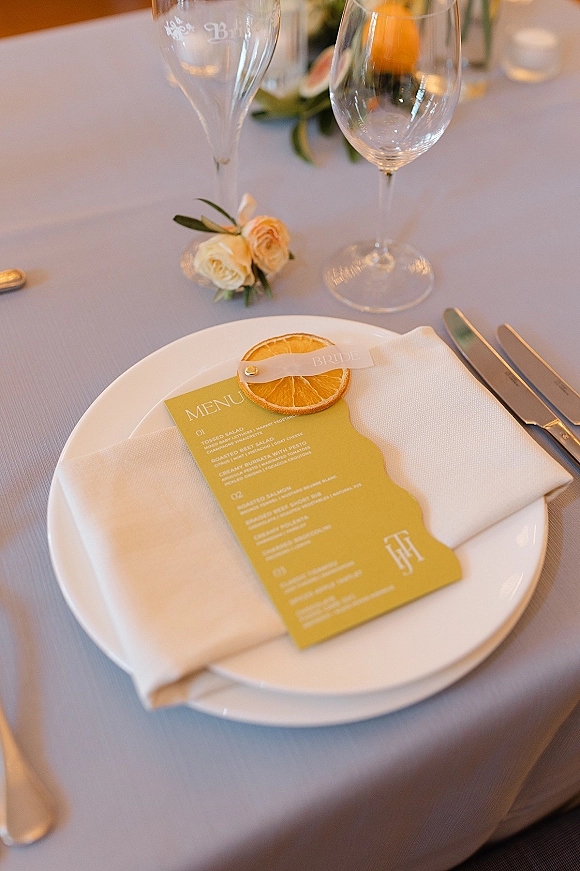 Wedding place setting with a wedding menu card and name tag on a white plate and napkin, with champagne flutes and candle on greenery tablecloth