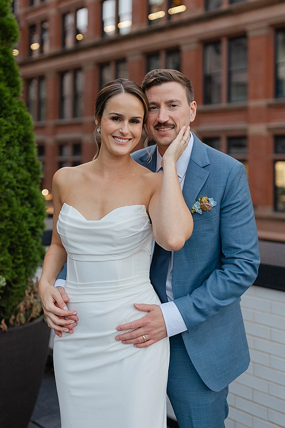 Couple portrait of bride and groom embrace, bride’s hand on groom’s face, in strapless dress and blue suit on a brick rooftop terrace