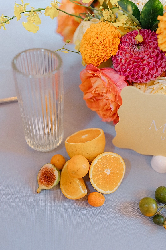 Wedding tablescape detail with a citrus wedding centerpiece of orange and pink flowers, oranges, kumquats, lime and fig in a ribbed vase on light linen