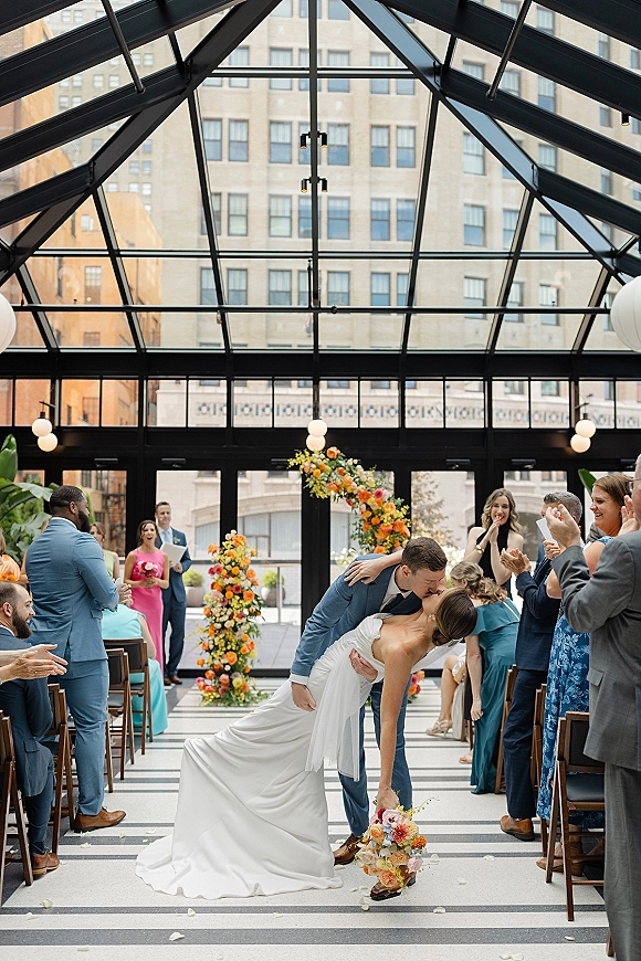 Wedding kiss portrait of bride and groom in a dip along a rose-petal aisle, with globe lights and a glass-ceiling atrium backdrop