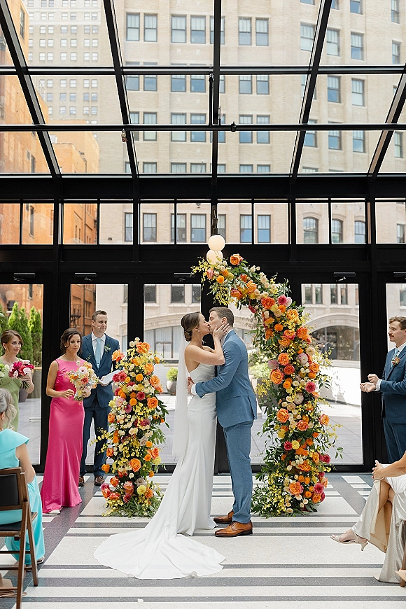 Wedding kiss portrait of bride and groom kissing beneath a colorful floral arch in a glass-ceiling atrium, bride in veil and strapless gown