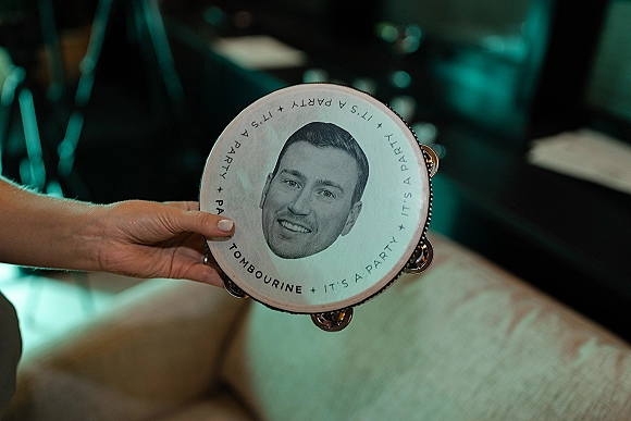 Wedding tambourine with a custom face print held in hand, jingles catching reception lighting beside chairs and table linens