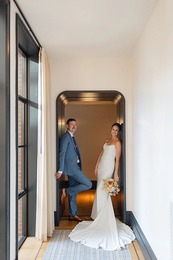Couple portrait of bride in a strapless wedding dress holding a bouquet beside groom in a blue suit in an indoor hallway by an arched doorway