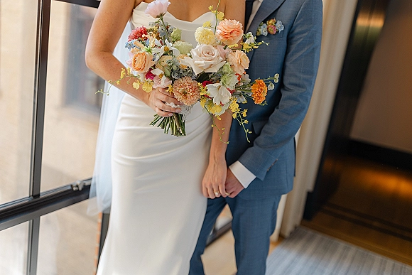 Wedding couple portrait with bride holding bouquet, standing in window light indoors as groom in blue suit holds her hand, rings visible