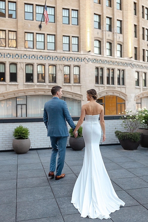 Couple portrait of bride and groom walking hand in hand, her strapless train gown and his blue suit on a city rooftop terrace