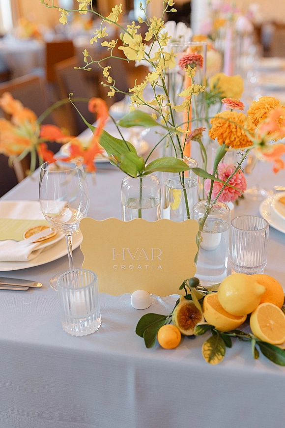Reception tablescape with wedding bud vase centerpieces of yellow orchids, bright flowers, and lemons on a white tablecloth with glasses and place card