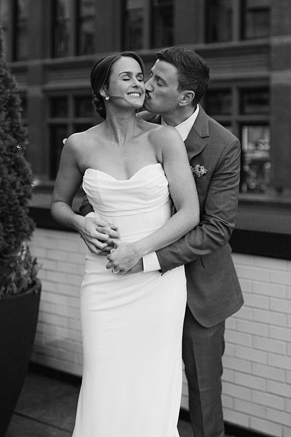 Wedding couple portrait of groom kissing bride’s cheek as they embrace in a strapless dress beside a brick wall and large windows