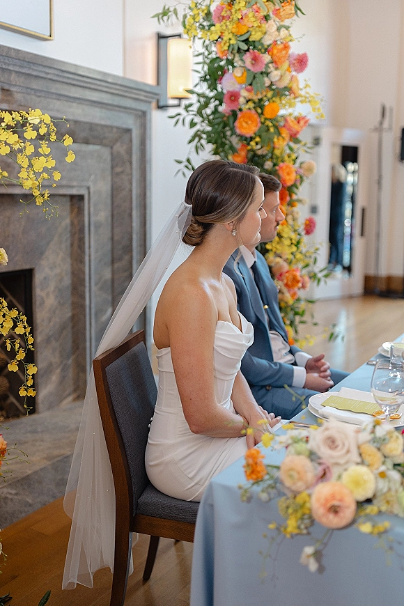 Wedding reception head table with bride and groom seated at head table, orange-pink floral centerpiece and blue tablecloth before a fireplace backdrop