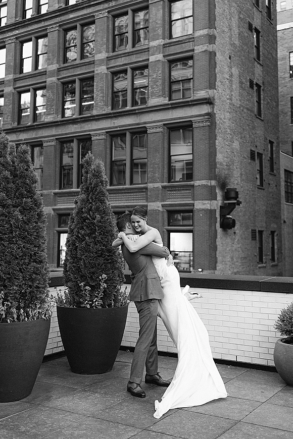 Couple portrait in a wedding hug photo as the groom lifts the bride in strapless dress and heels on a brick rooftop terrace with planters