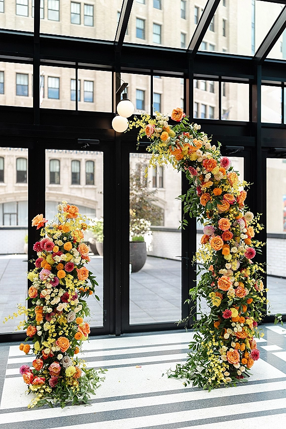 Floral wedding arch with an asymmetrical spray of roses, mixed florals, and greenery framing glass doors in a modern venue
