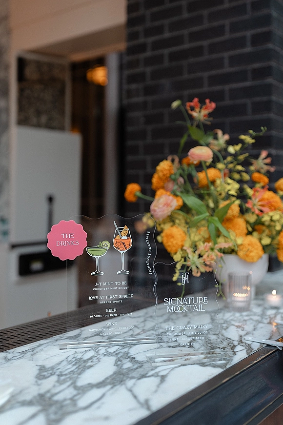 Wedding bar signage on a marble bar top with acrylic drink menu sign, cocktail illustrations, orange florals, and votive candles by a brick wall