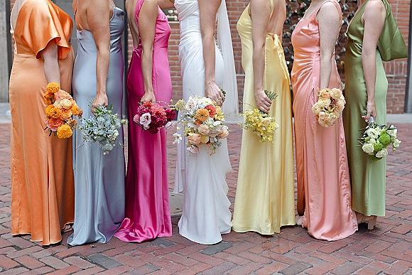 Bridesmaid dresses in mismatched bridesmaid dresses line up on a brick street, holding bouquets, wearing satin slip styles with side slits and heels