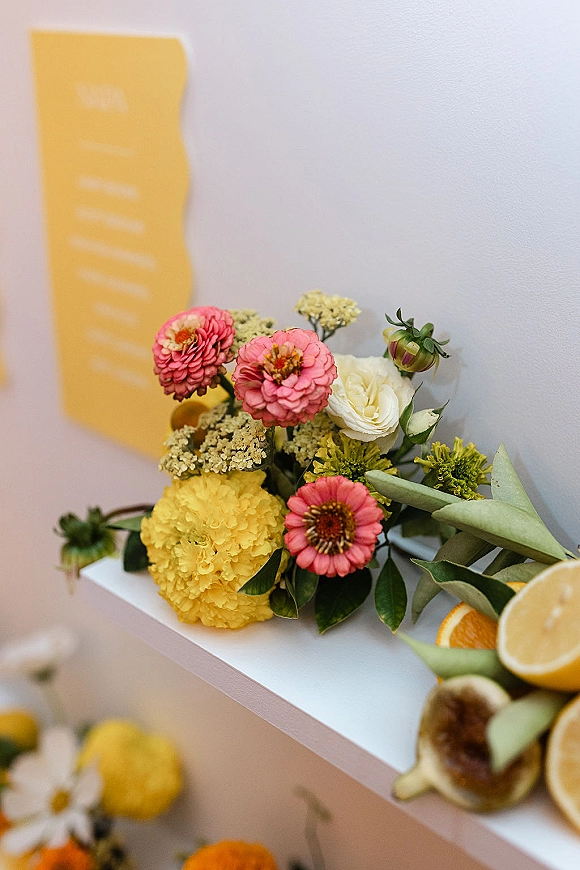 Wedding bouquet with colorful wedding bouquet blooms, greenery, and citrus fruit beside a place card on a tabletop against a white wall
