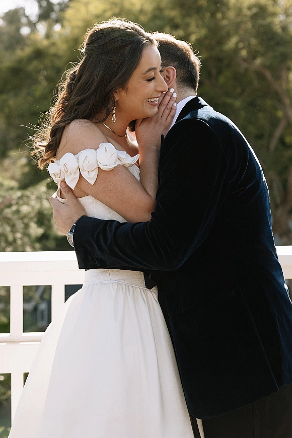 Wedding couple portrait with bride and groom embrace, groom whispering as they lean forehead to forehead by trees and a white railing