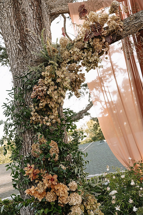 Wedding floral installation with a floral garland of greenery, hydrangeas, orchids and anthurium draped over tree branches in a garden setting