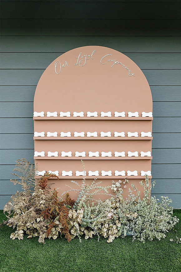 Wedding escort display with an arched escort card wall and dog bone place cards, calligraphy sign, and floral ground arrangement on grass lawn