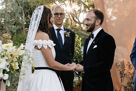 Wedding vows as bride and groom hold hands, her lace veil and off-shoulder dress backlit at a sunny outdoor garden ceremony