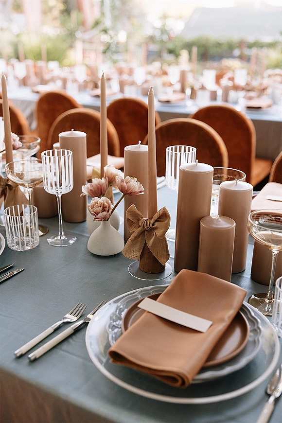 Reception tablescape with wedding table setting of taper and pillar candles, layered plates, striped glassware, and bud vase on long outdoor tables in soft daylight