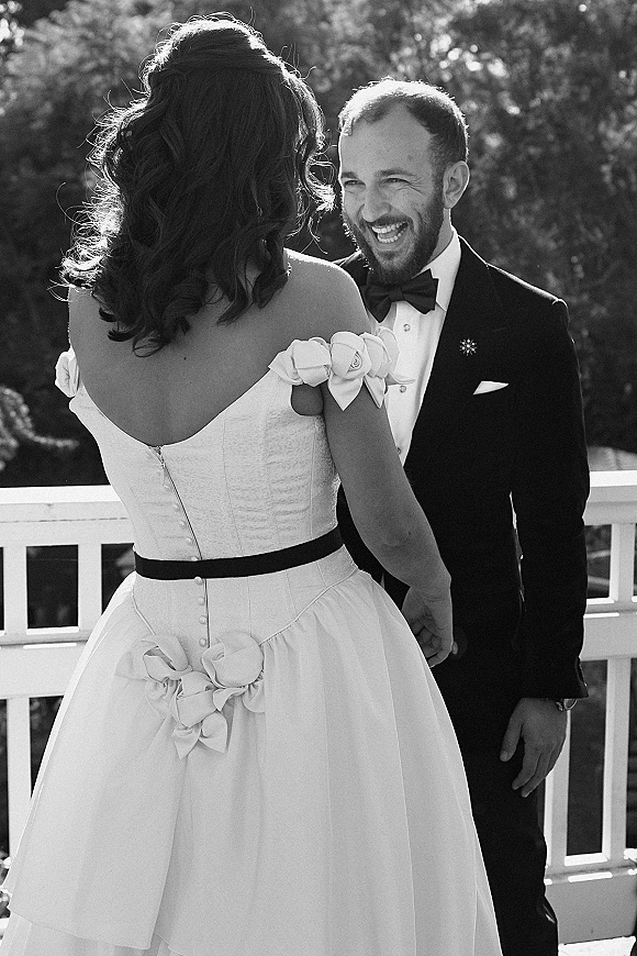 Couple portrait of bride and groom laughing while holding hands on an outdoor terrace, her strapless gown with bow sleeves and black sash