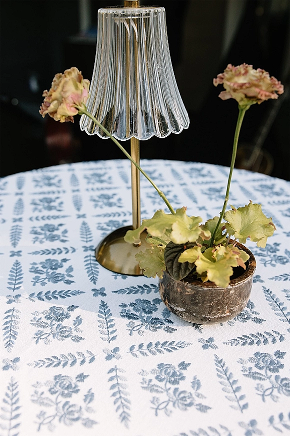 Wedding table decor with a reception table centerpiece featuring a small brass lamp, pleated glass shade, bud vase and flowers on patterned cloth