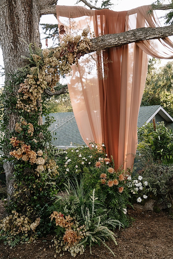 Wedding ceremony backdrop with draped fabric and an asymmetrical floral installation on a tree, greenery garland and grounded florals in a garden setting