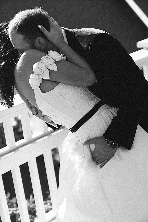Wedding kiss portrait in black and white as groom dips the bride in a strapless gown with sash, her hand on his head by a white railing