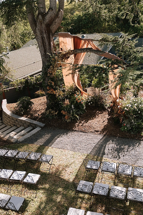 Ceremony setup with outdoor wedding ceremony seating, peach draped fabric and asymmetrical floral arch beneath a large tree in sunlight