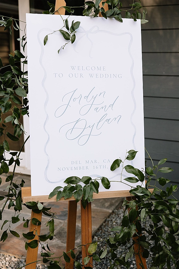 Wedding welcome sign with calligraphy lettering on a wood easel, framed by greenery garland beside house siding and stone walkway