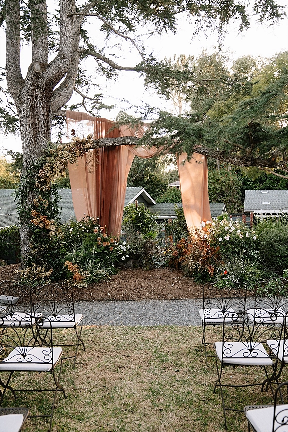 Ceremony setup with outdoor wedding ceremony seating, floral arch on a large tree and terracotta draped backdrop on a garden lawn