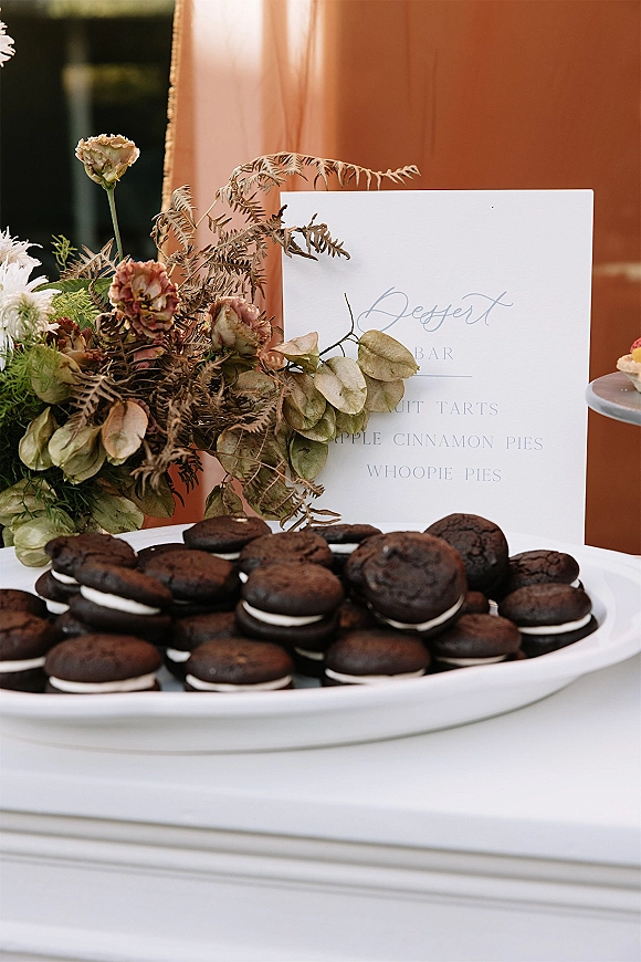 Dessert table with wedding dessert table sign and whoopie pies on a platter, styled with dried greenery against a terracotta backdrop