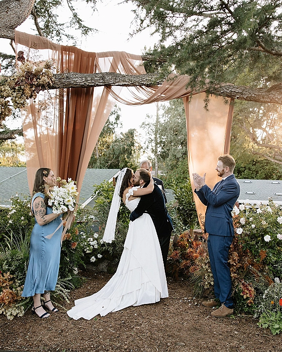 Ceremony kiss under a draped wedding arch with florals as the bride in a veil and groom in a black suit embrace on a rooftop garden