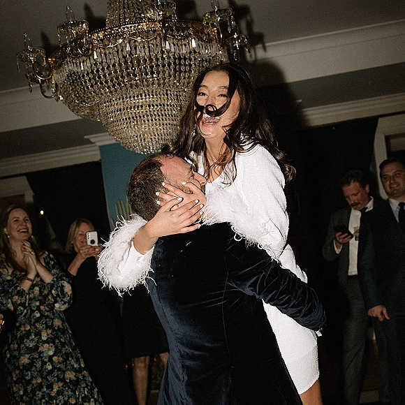 Wedding reception dance as groom lifts laughing bride in a feather-sleeve mini dress under chandelier, guests filming in dim ballroom