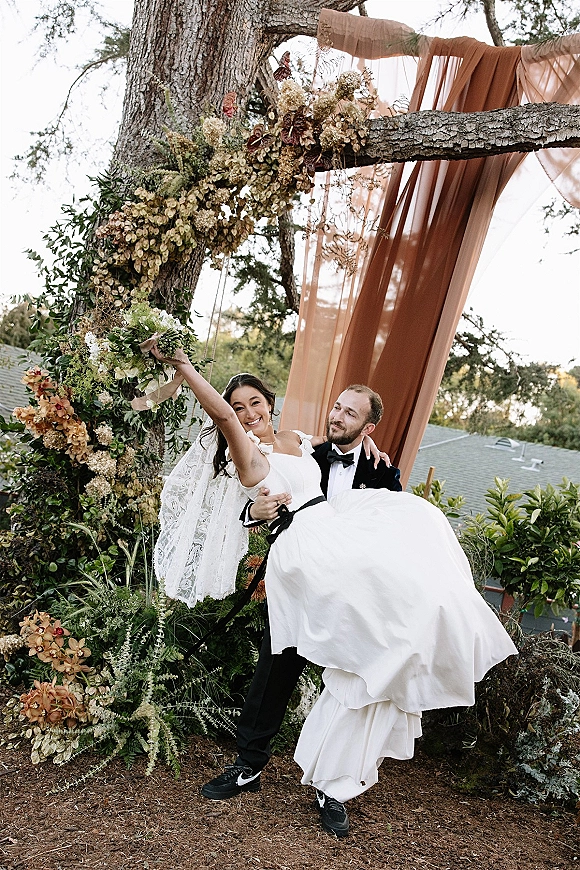 Couple portrait of a groom carrying the bride in a wedding dip pose beneath a floral tree arch on a rooftop garden, sky behind
