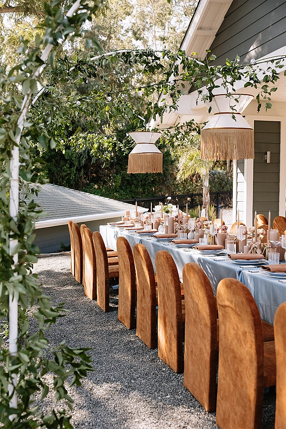 Reception tablescape with light blue tablecloth on a long outdoor reception table, candles and bud vases under string lights on a patio