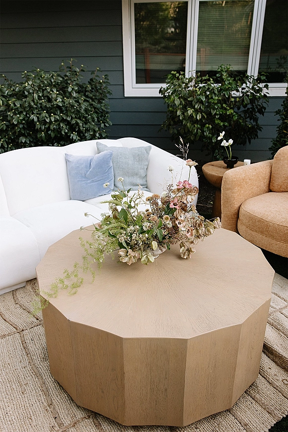 Wedding lounge decor with outdoor sofa, throw pillows, and low coffee table topped with floral arrangement beside house siding and shrubs