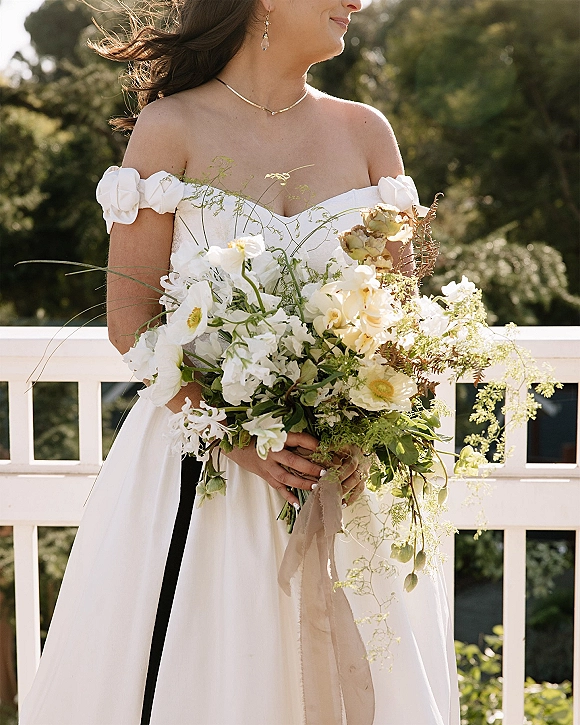 Bridal portrait of a bride holding bouquet, wearing an off the shoulder wedding dress with white blooms and greenery, sunlight by railing