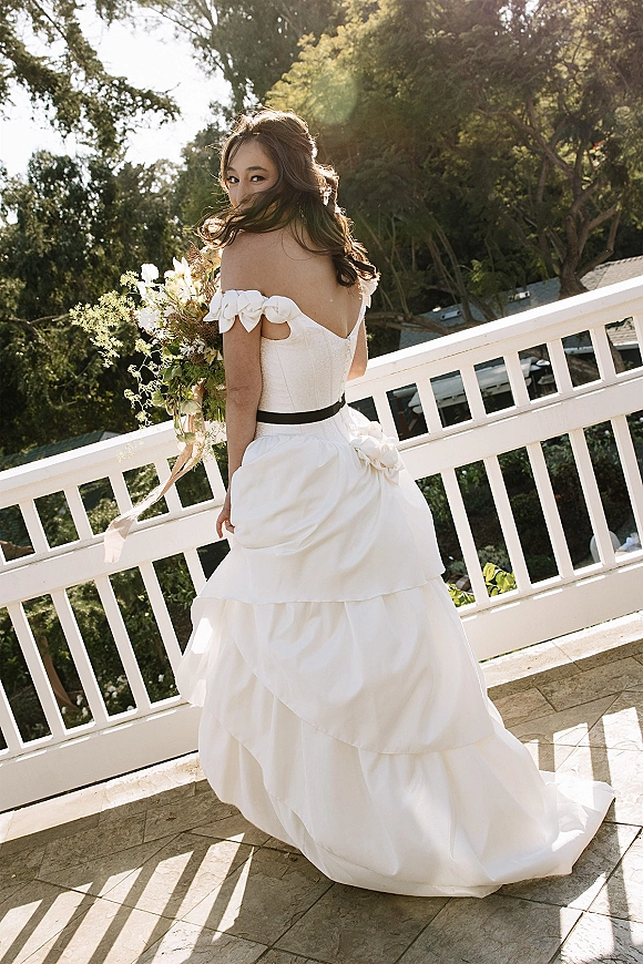 Bridal portrait of a bride in an off the shoulder wedding dress, looking back on a sunlit balcony, holding a bouquet with greenery.
