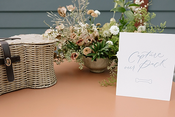 Wedding photo guestbook station with a wicker picnic basket, floral arrangement in a ceramic vase, and calligraphy sign on a peach tabletop