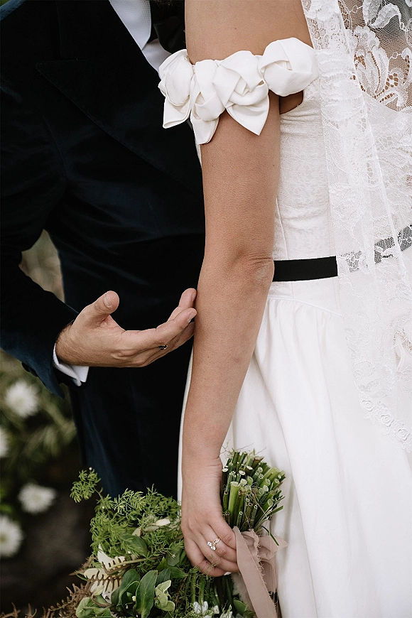 Bridal bouquet of lush greenery wedding bouquet held against a bride’s lace veil and black sash, with groom’s hand and ring in view