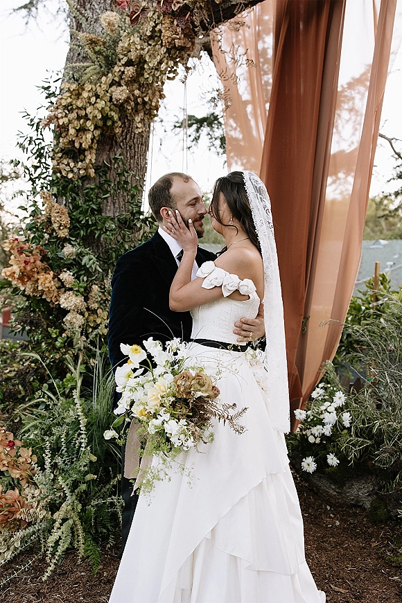 Wedding kiss portrait of bride and groom kissing, bride holding his face in lace veil beside a floral arch with greenery outdoors